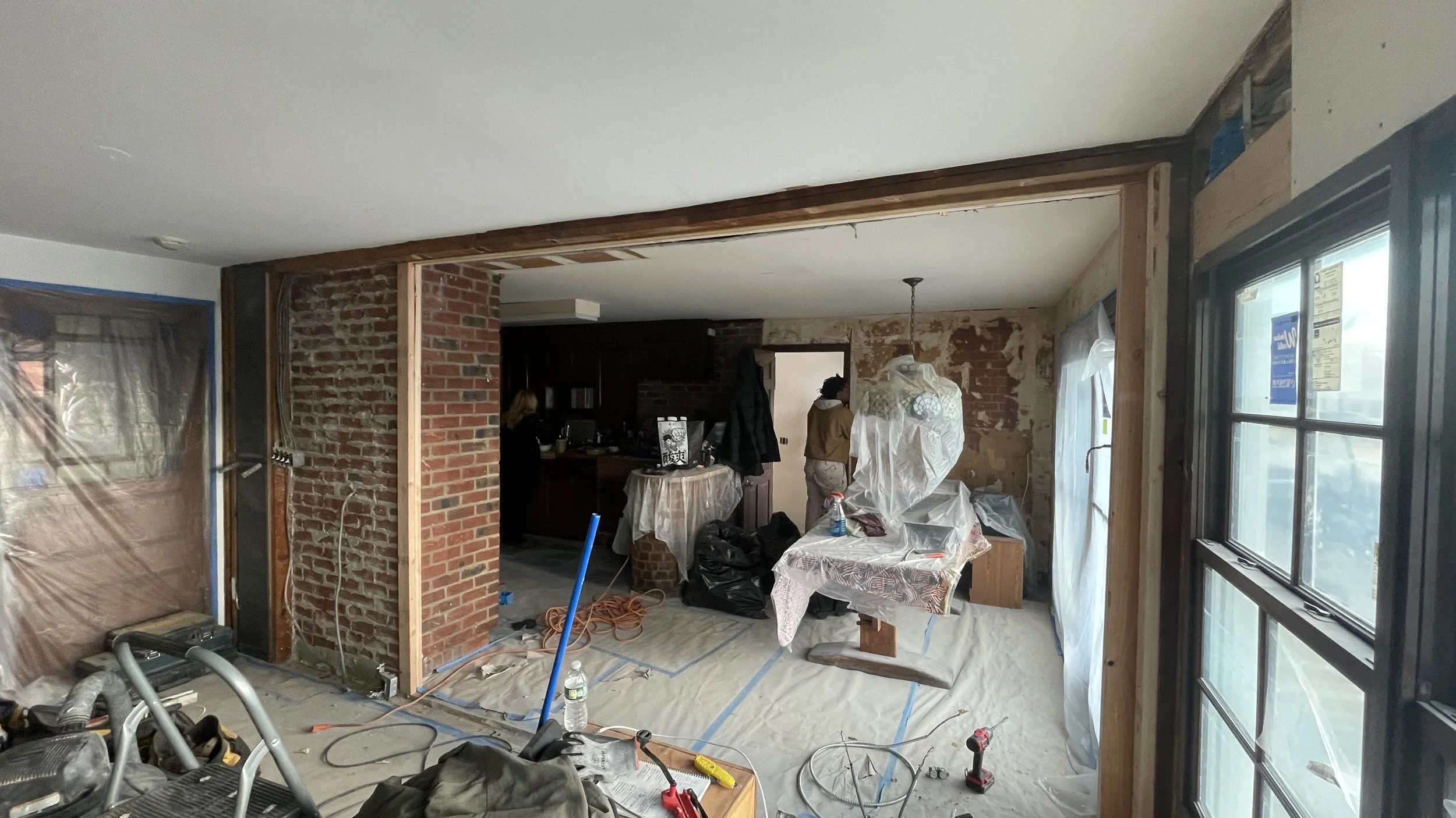 Living room undergoing renovation with exposed brick and drywall, covered windows, construction tools, and two workers.