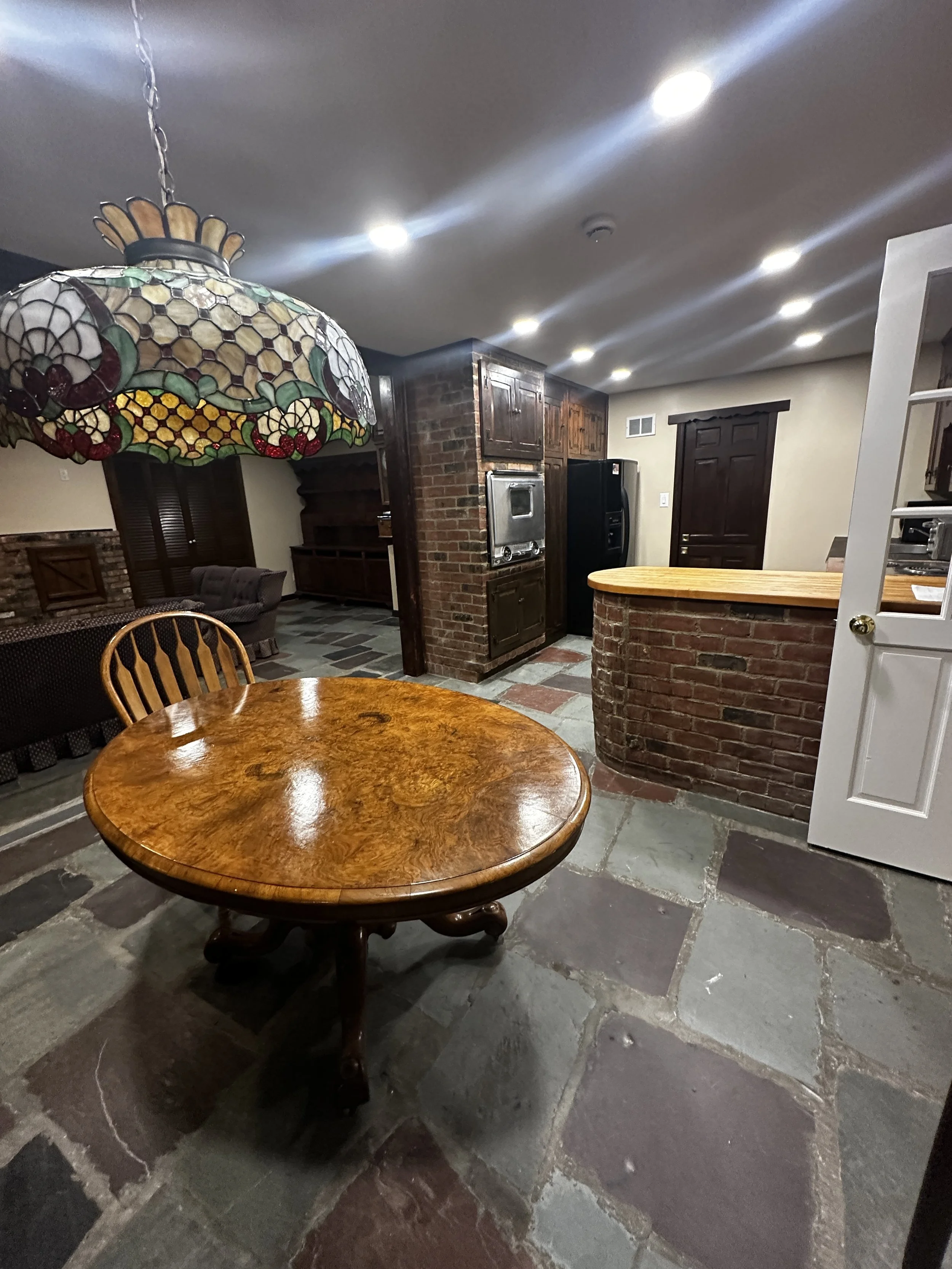 Interior of a house with a wooden table, stained glass hanging lamp, brick columns, dark wood cabinets, black refrigerator, and a closed dark wood door. The floor is made of large stone tiles.