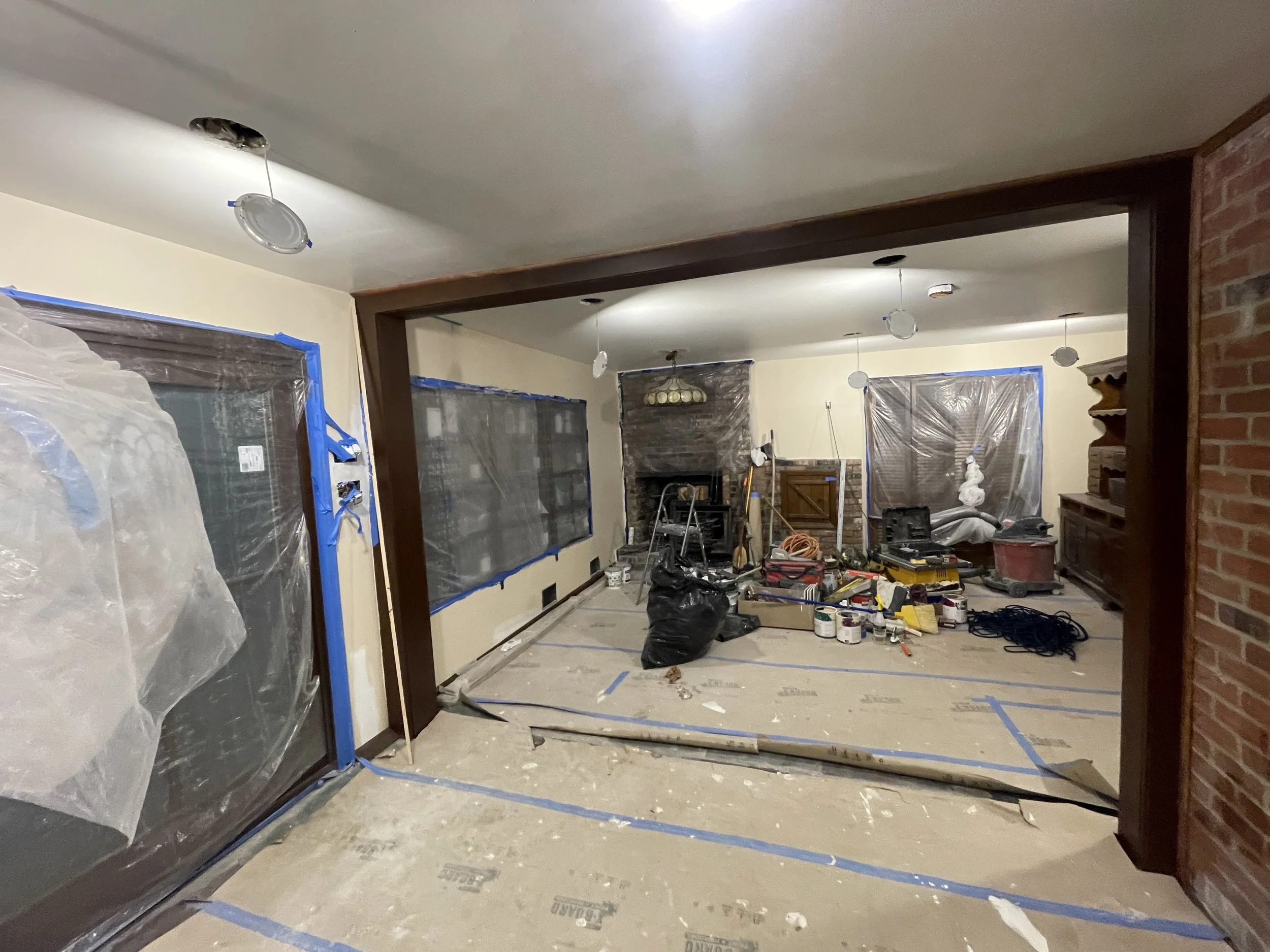 Living room under renovation with plastic-covered furniture, ceiling lights, construction tools, and supplies scattered across the floor.
