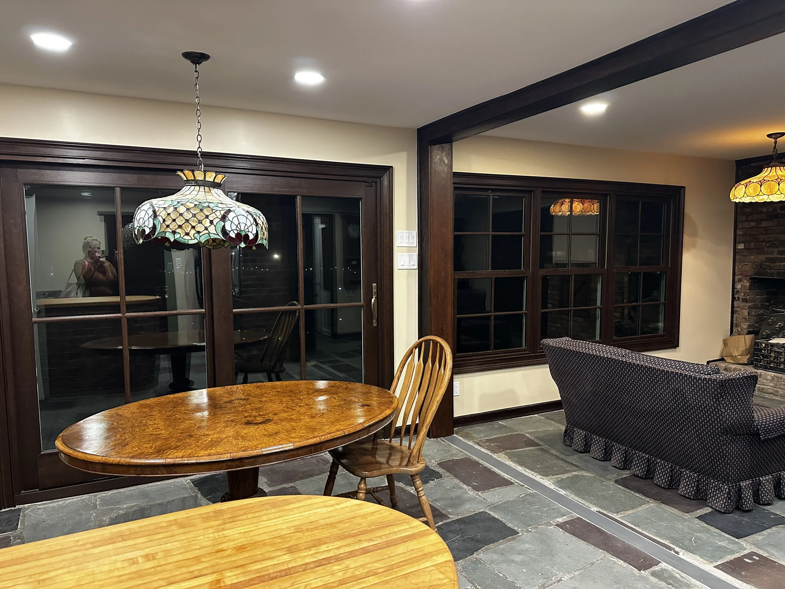 A dining room with a stained glass Tiffany-style pendant light hanging over a wooden round table with matching chairs. Large wooden-framed glass doors lead to a dark outdoor area. To the right, a section of a brick fireplace and a window with multipl