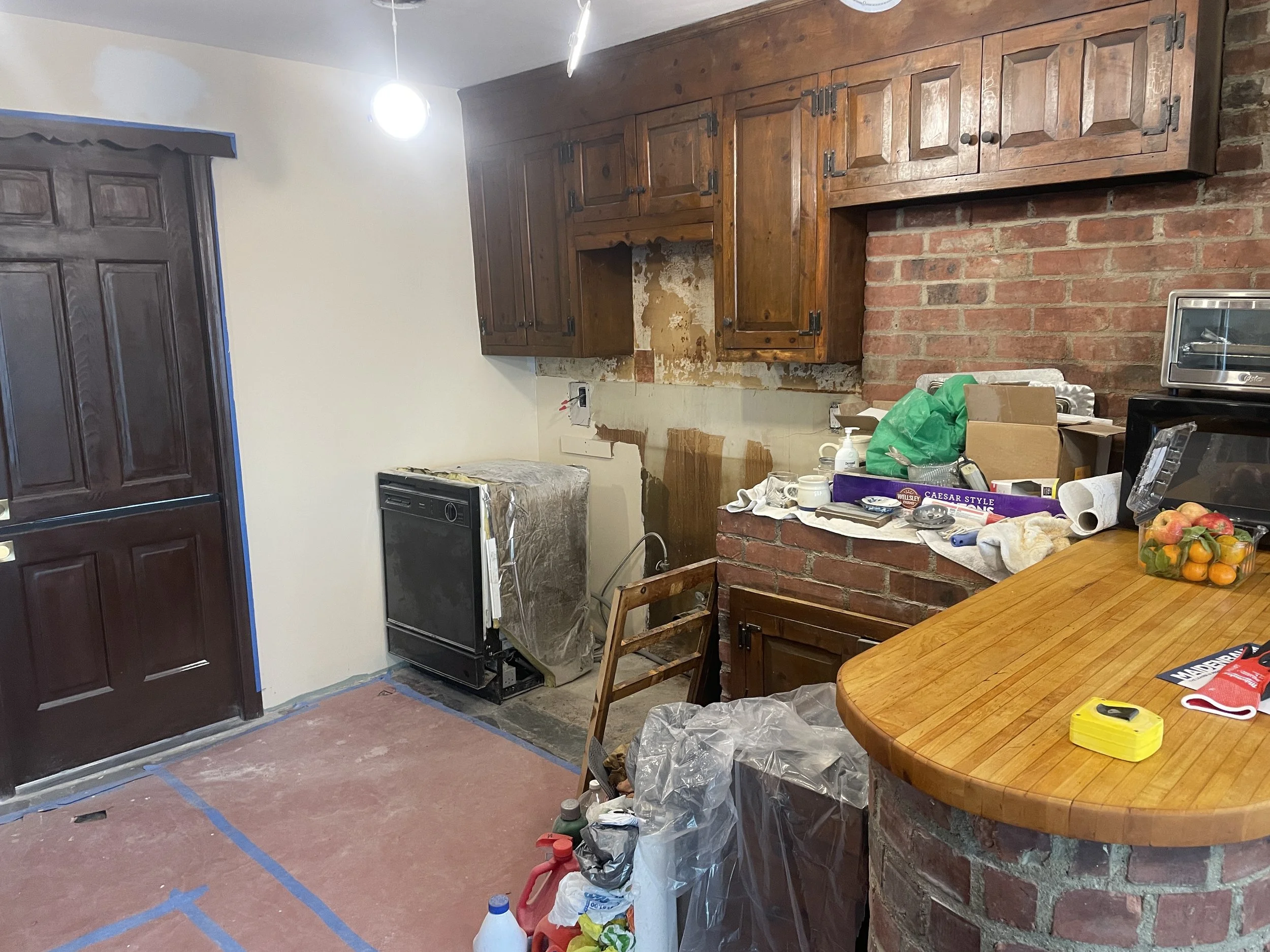 Kitchen under renovation with unfinished wall and cabinet removal, construction materials on countertop, and painting supplies on table.