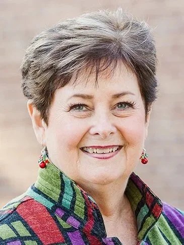A smiling woman with short gray hair, wearing colorful earrings and a multicolored jacket.