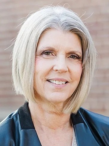 A portrait of a smiling middle-aged woman with shoulder-length gray hair, wearing a black jacket, standing outdoors in front of a brick wall.