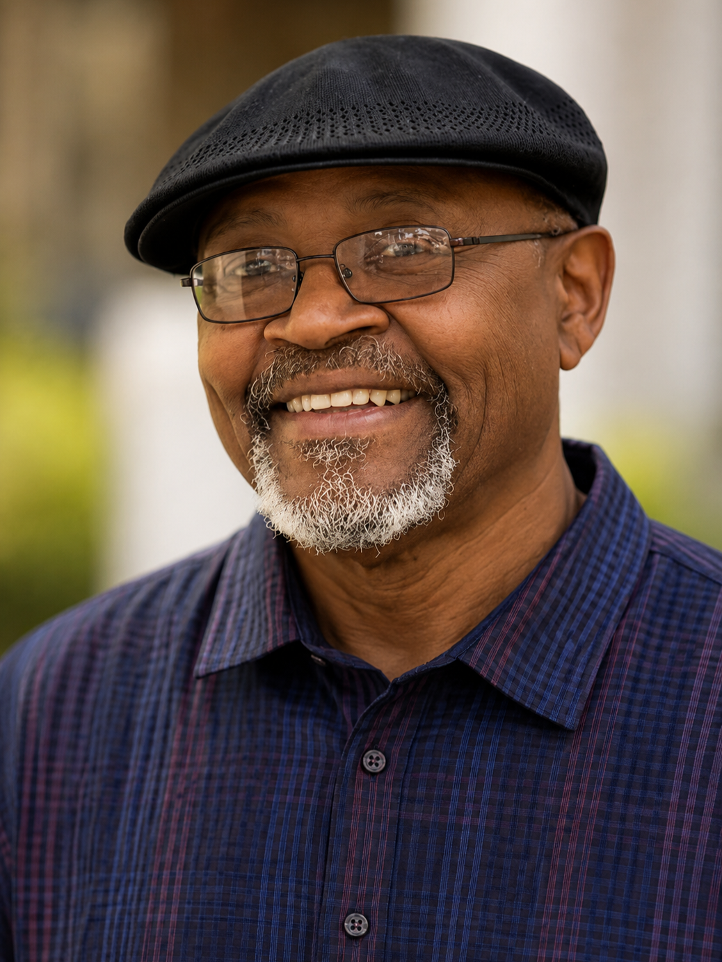 Close-up of an older African American man wearing glasses, a black cap, a blue checkered shirt, smiling outdoors with a blurred background.