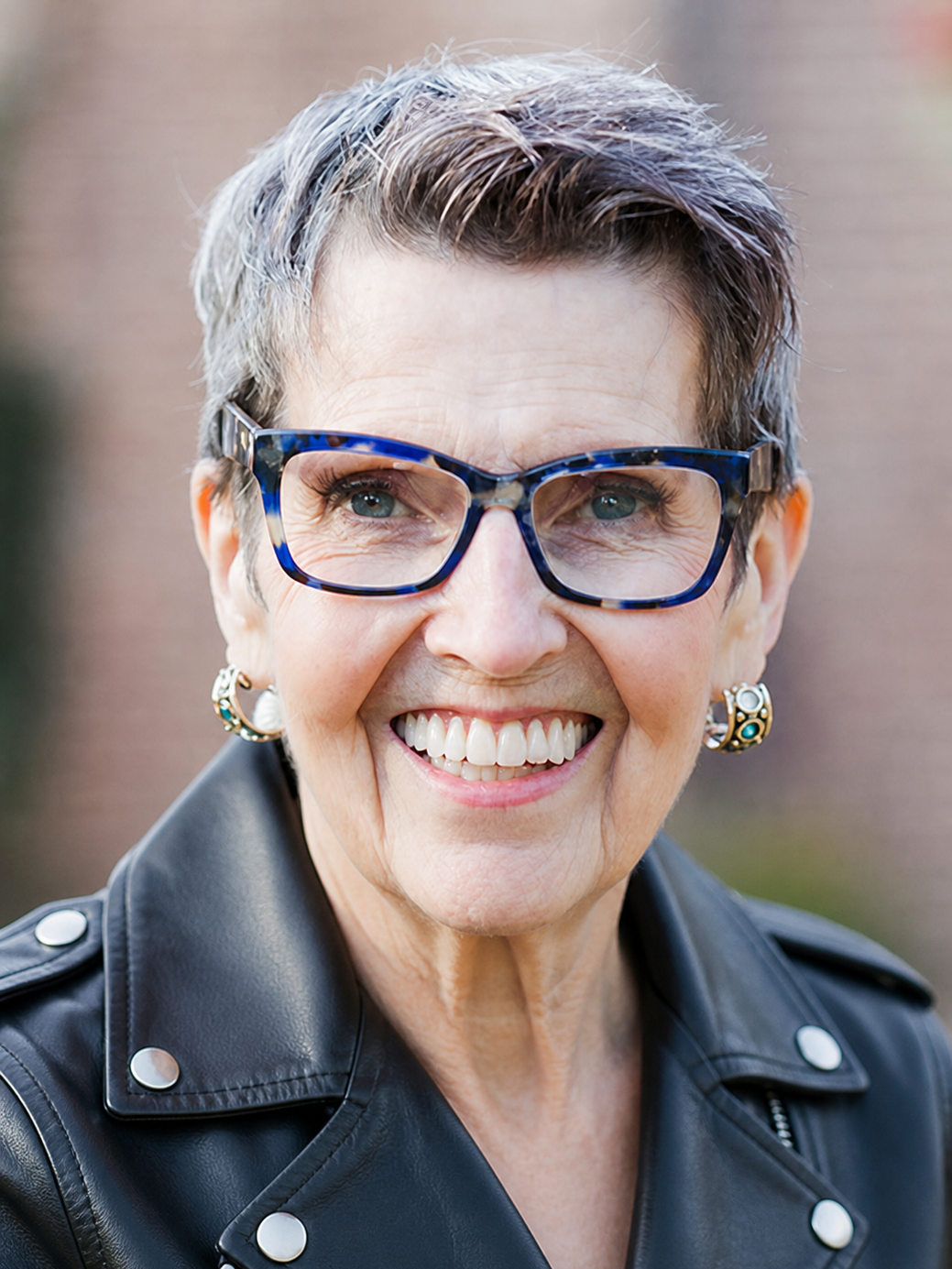 Close-up of a smiling woman with short gray hair, wearing blue glasses, statement earrings, and a black leather jacket.