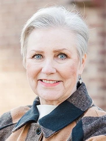 Close-up of an older woman with short gray hair, blue eyes, and a warm smile, wearing a brown and black checkered coat.