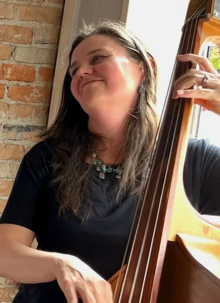 Woman with long hair and a necklace playing a double bass, smiling and looking to the side, with a brick wall in the background.