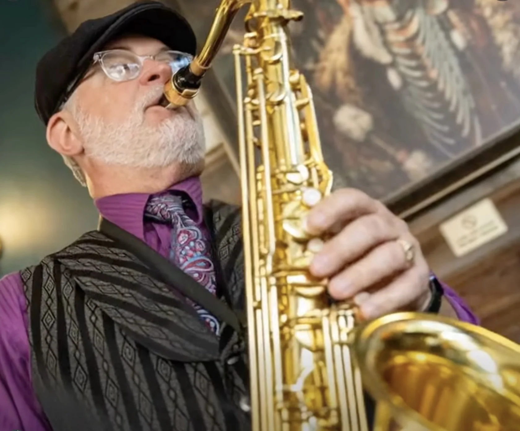 An older man with glasses, wearing a cap and a pinstriped shirt with a patterned tie, playing a saxophone with closed eyes in a dimly lit setting.