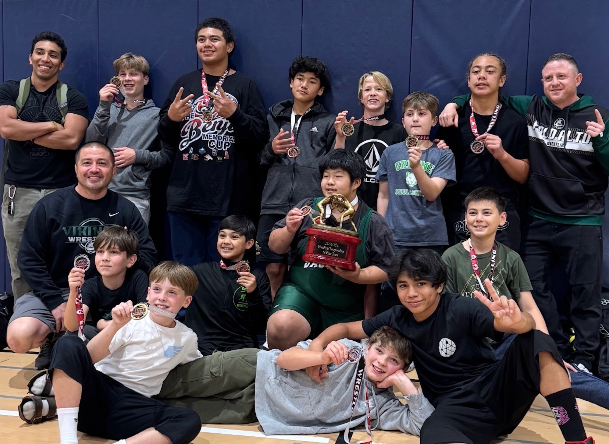 Group of young wrestlers with medals and a trophy, smiling and posing after a competition in a gymnasium.