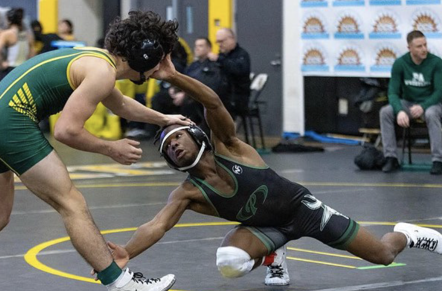 Two male wrestlers compete on a mat during a match, with one wrestler in green and yellow and the other in black and green, while referees and spectators watch in the background.