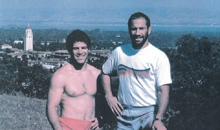 Two men standing outdoors with a city skyline in the background. One man is shirtless with curly hair, the other is wearing a white T-shirt and gray shorts.