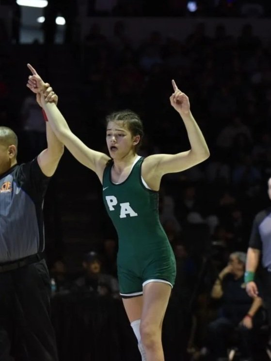 A young female athlete in a green uniform with white letters 'PA' celebrating after a sports event, with her arms raised and pointing upwards.