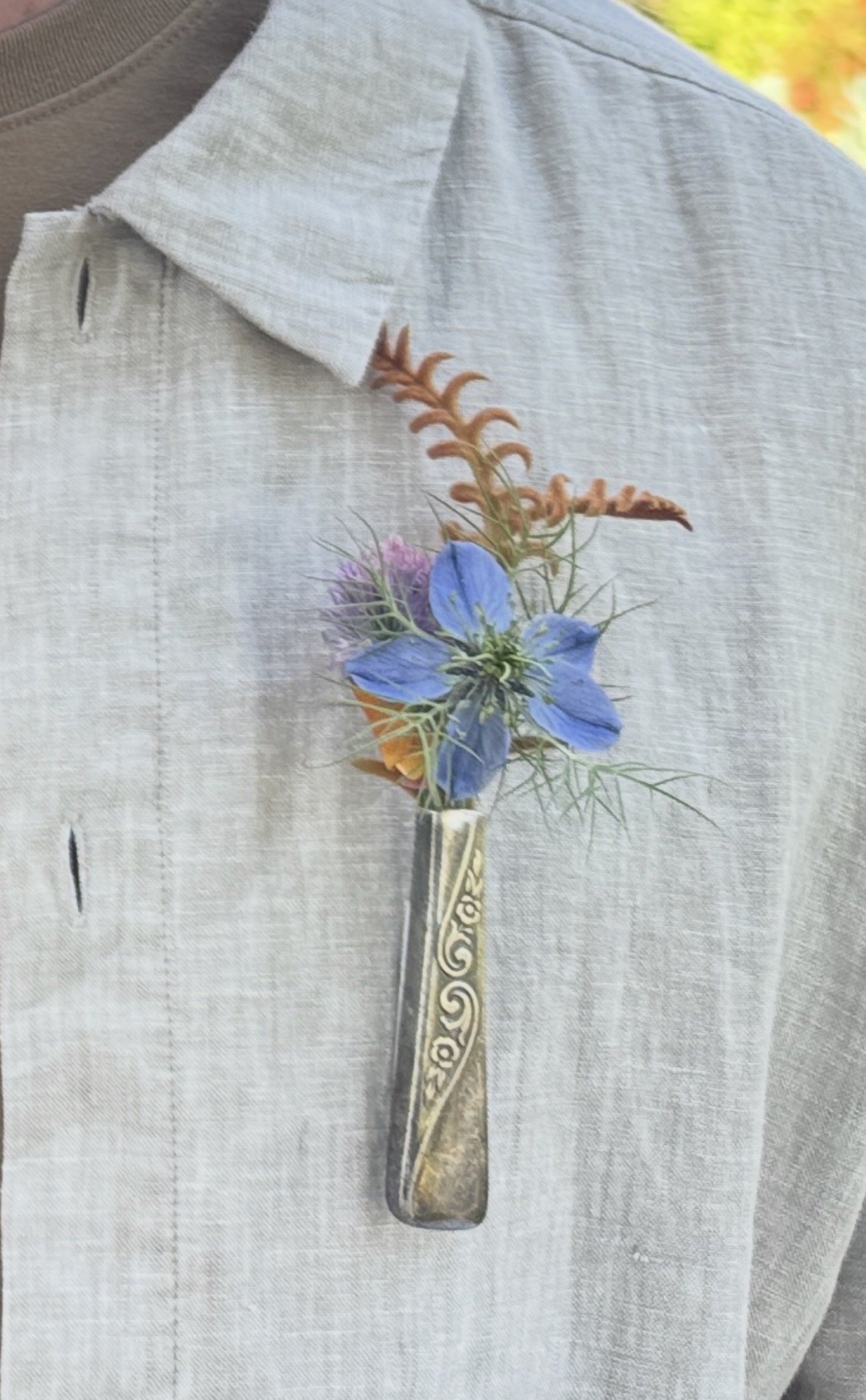 A close-up of a person wearing a light gray shirt with a floral boutonniere attached to the shirt pocket, featuring blue flowers and orange accent flowers. The shirt fabric is textured and the person's arm is also visible.