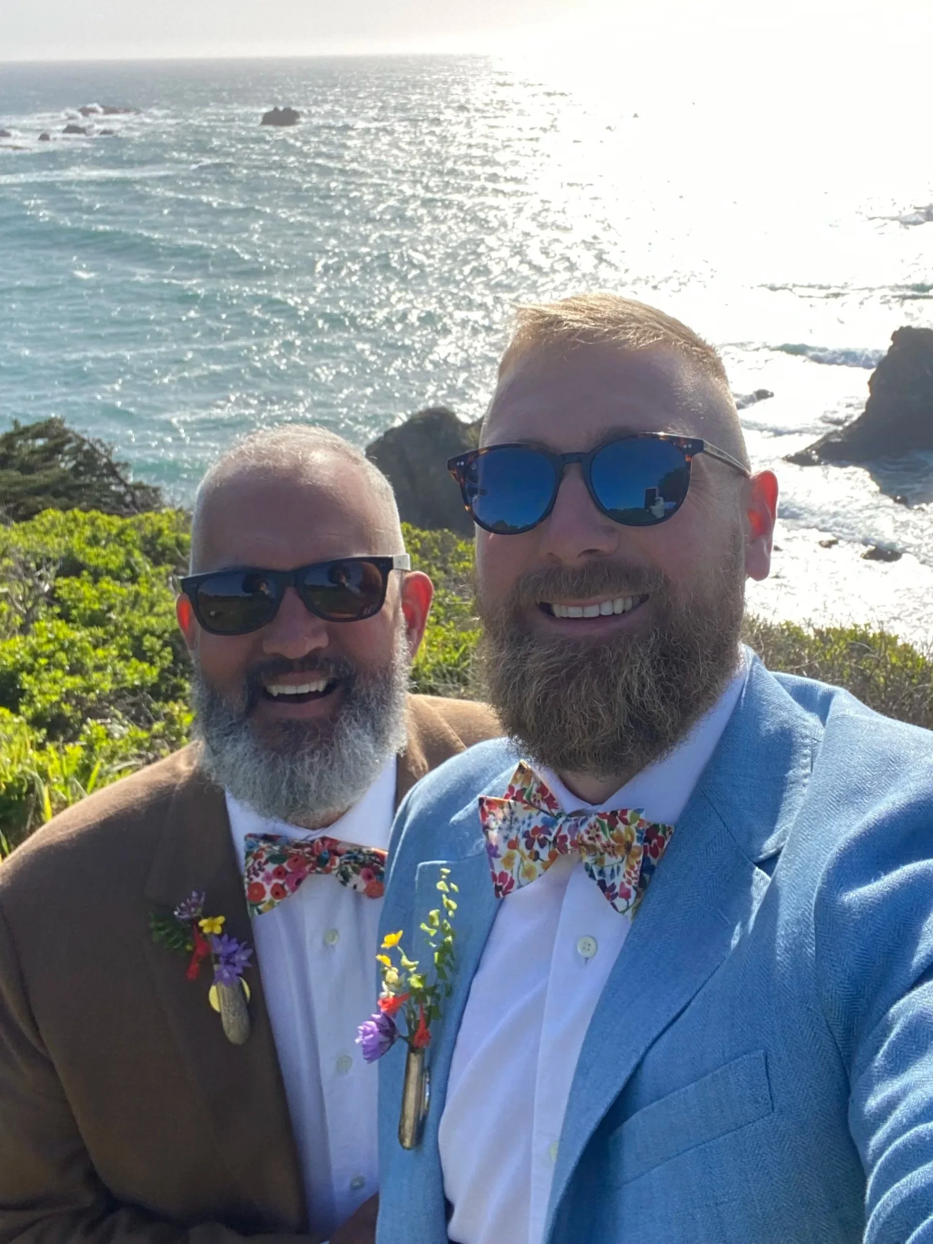 Two men dressed in suits, wearing colorful bow ties and sunglasses, smiling for a photo near a rocky coastline with the ocean in the background.