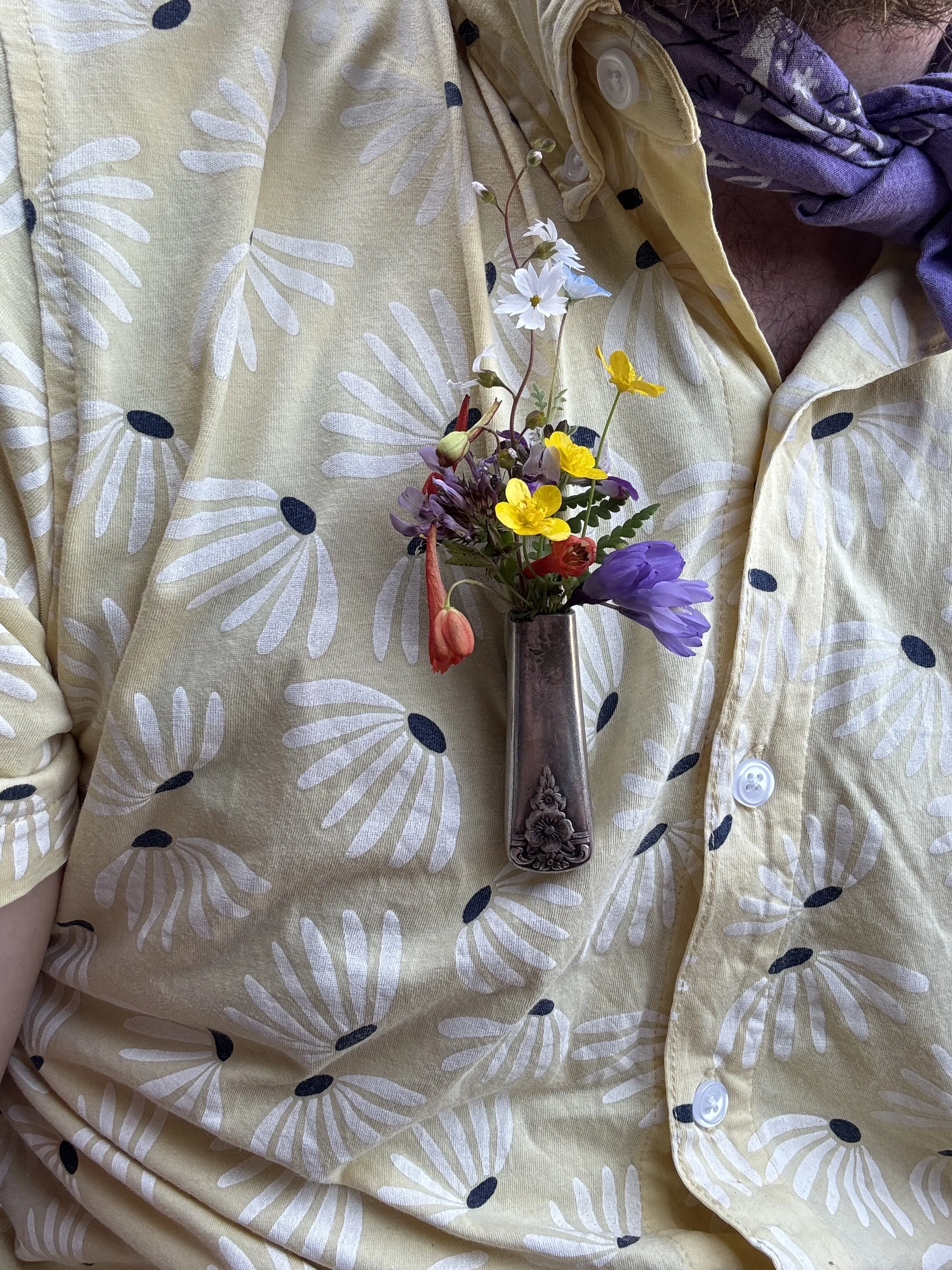A close-up of a person wearing a yellow button-up shirt with a floral pattern, with a small bouquet of colorful flowers in front of their chest.