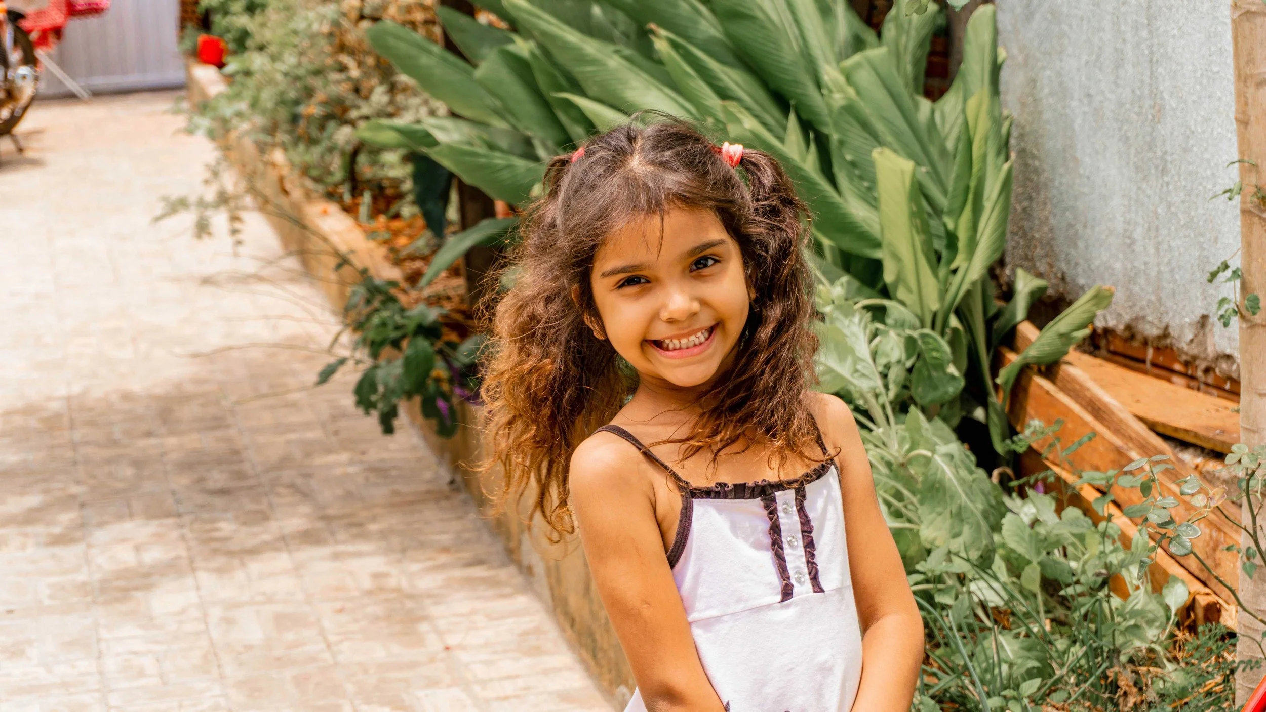 A young girl with curly brown hair smiling and standing outdoors in front of green plants.
