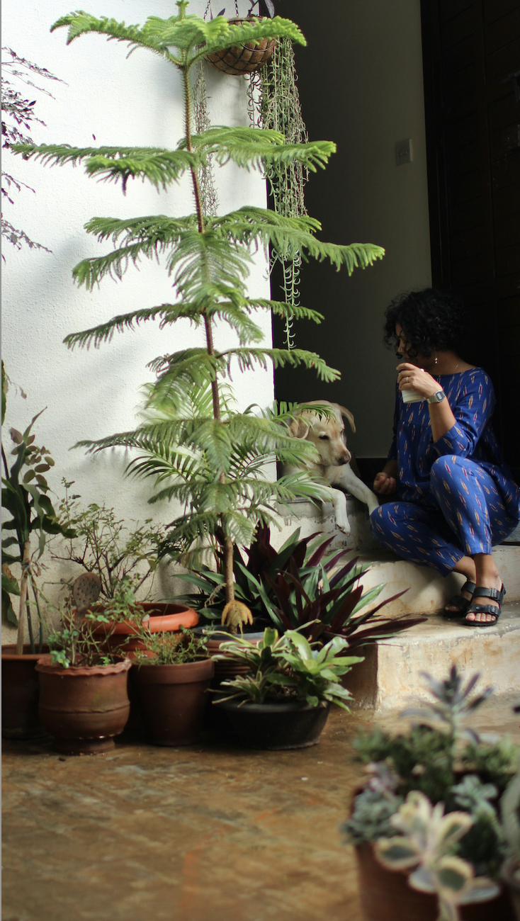A woman in blue pajamas with a dog resting beside her on a step, surrounded by potted plants on the ground, with a white wall in the background.