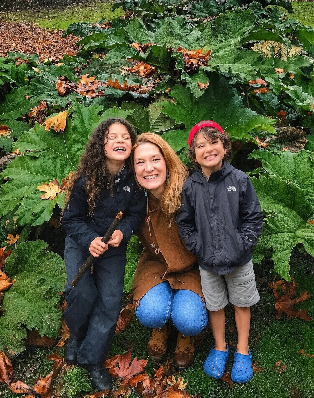 A woman with two children smiling and posing outdoors amongst large green and brown autumn leaves.