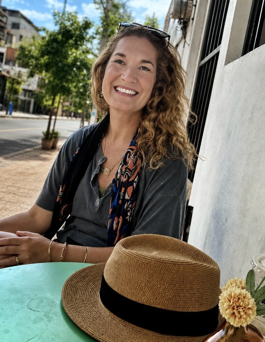 A woman with curly hair and sunglasses on her head smiling while sitting outdoors at a table. She is wearing a gray shirt, a colorful scarf, and jewelry. There is a straw hat and a small bouquet of flowers on the table in front of her.