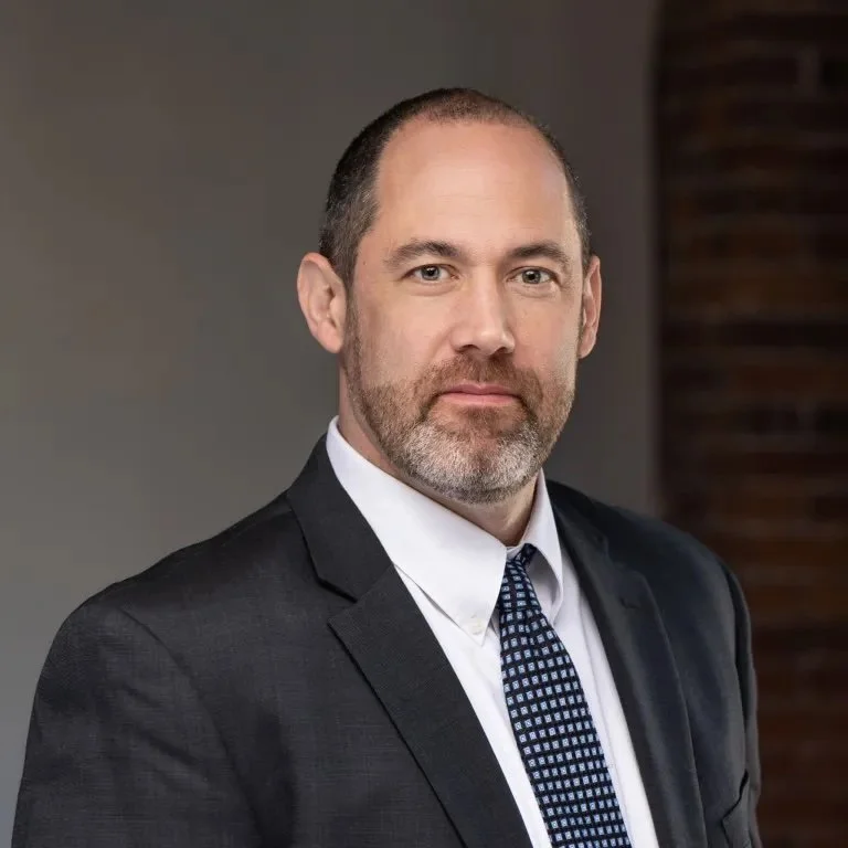 A professional man with short hair and a beard, wearing a dark suit, white shirt, and patterned tie, looking at the camera in a formal setting.
