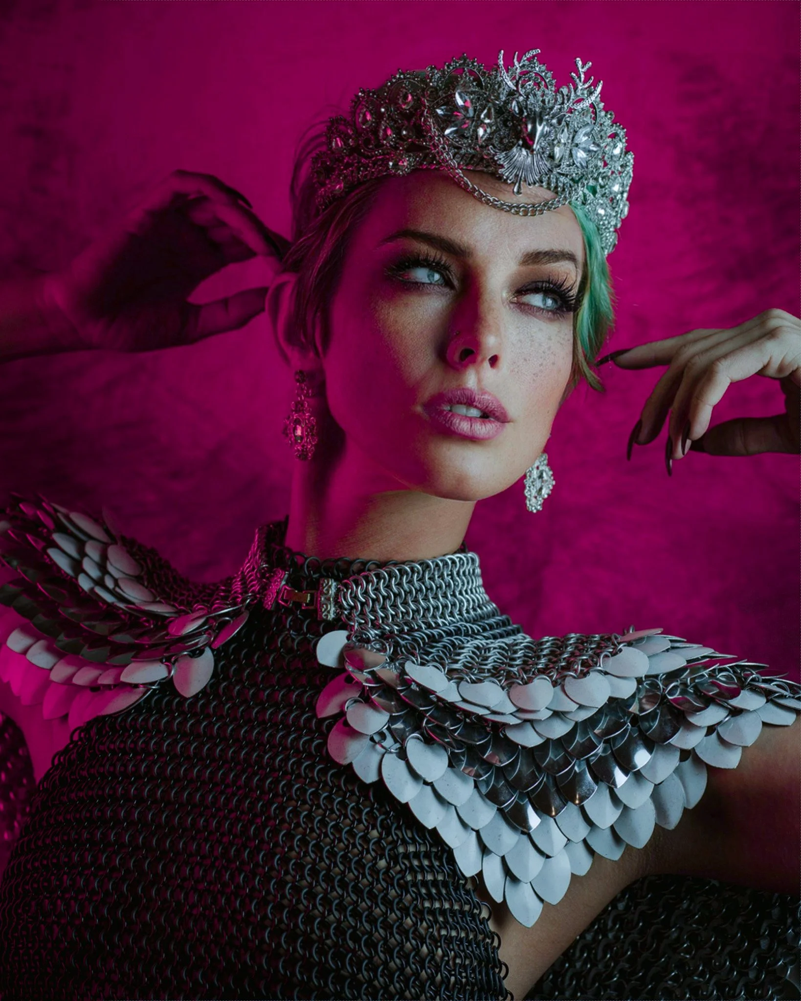 Close-up of a woman with makeup, green hair, wearing a silver tiara, earrings, and a necklace, with a pink background and metallic armor-like accessories.