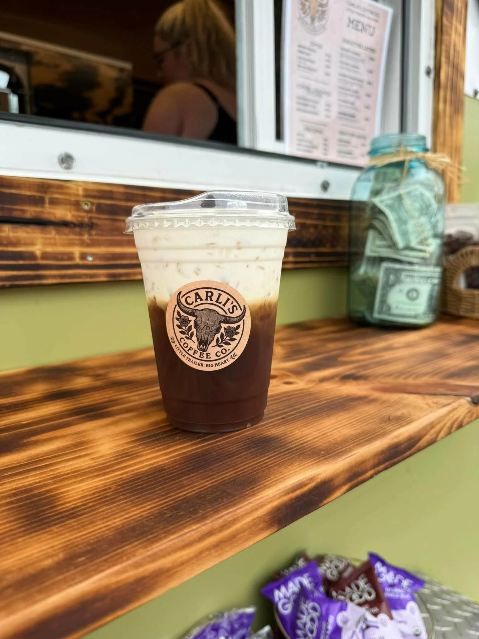 A plastic cup of iced coffee with a logo on the front sits on a wooden counter. In the background, a woman with blonde hair and glasses is visible through a window, along with a menu and a jar of cash. Part of a purple snack bag is also visible in the foreground.