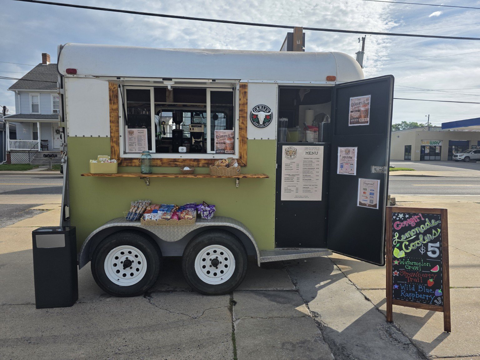 A green and white mobile coffee trailer parked on the street with a front window, open door, and colorful chalkboard sign advertising flavored Lemonade Coolers for $5 in front.