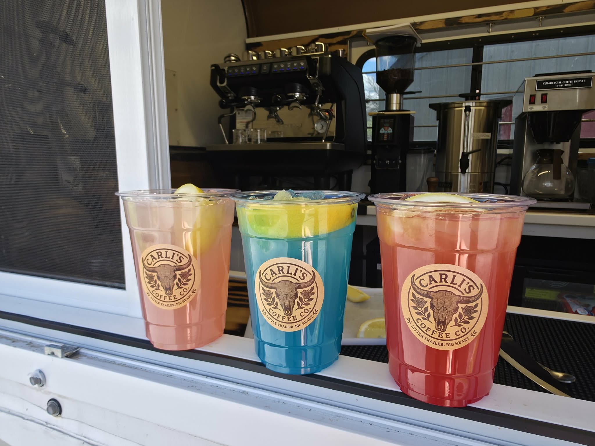Three colorful drinks in cups with a logo of Carl's Coffee Co. inside a coffee stand, with a coffee machine and brewing equipment in the background.