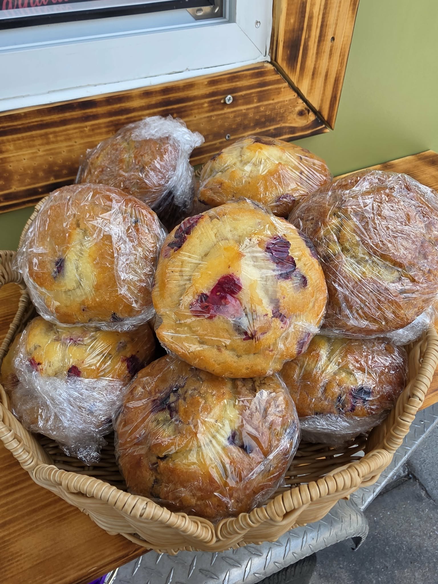 A basket of wrapped muffins with visible fruit fillings, placed on a wooden surface near a green wall and a bulletin board.