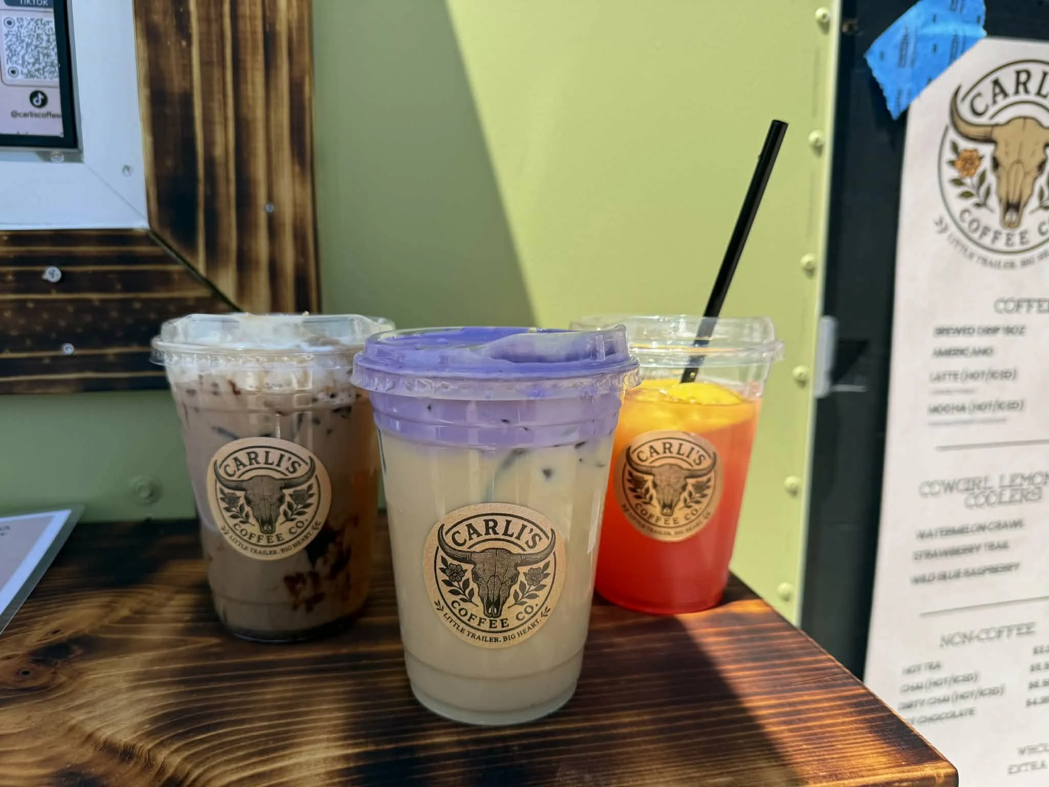 Three iced beverages in plastic cups with black straws, on a wooden counter at a coffee shop, branded with a logo featuring a cow skull and the name 'Carl's Coffee Co.'.