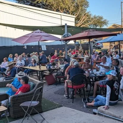 Crowd of people dining outdoors at a restaurant with umbrellas, with some sitting at tables and others on benches, under clear blue sky.