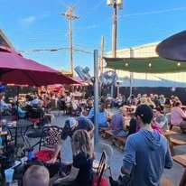 Crowd gathered outdoors, some sitting at tables under umbrellas, at a lively event or festival.