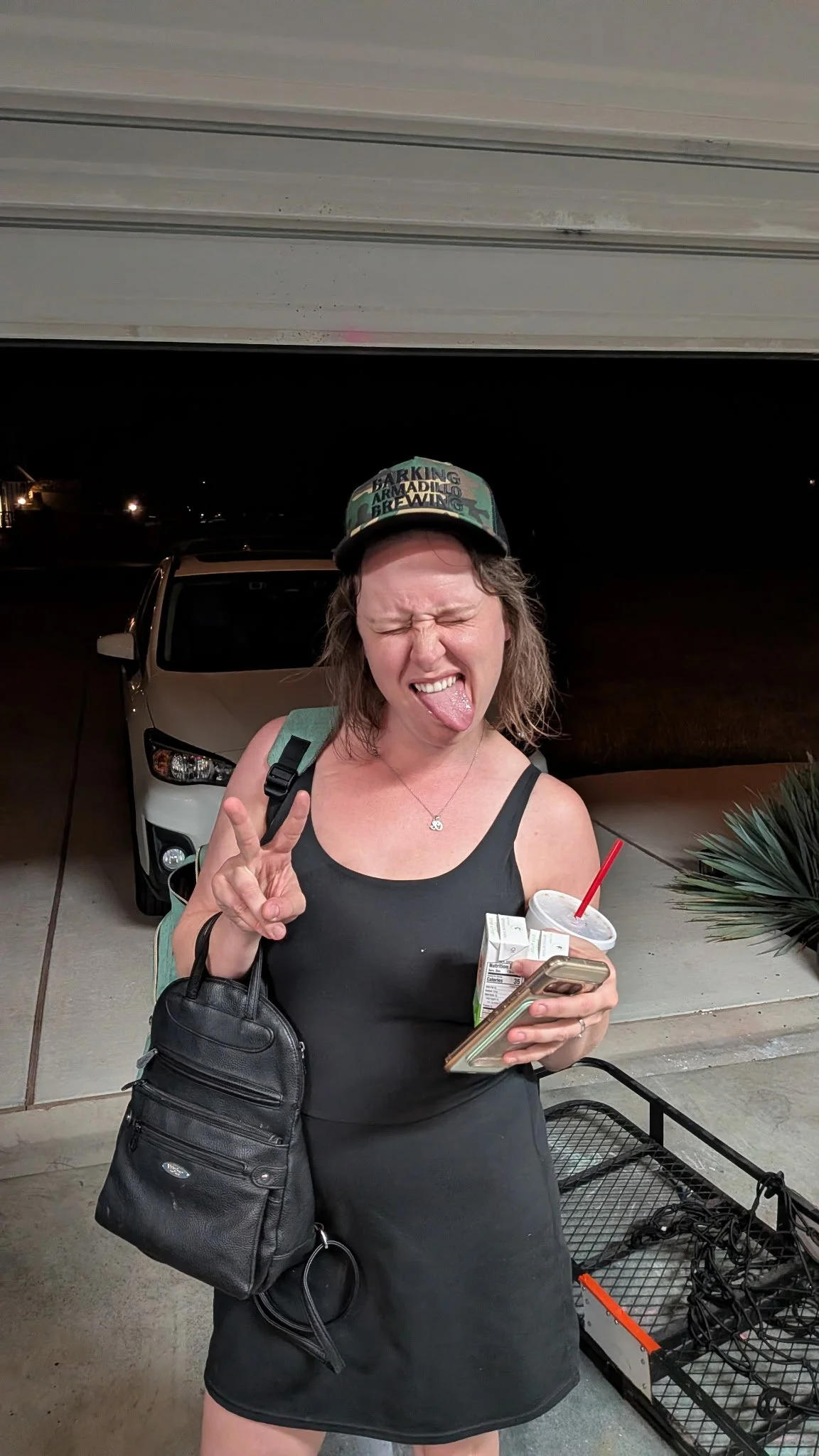 A woman making a peace sign with her right hand, sticking out her tongue, wearing a black sleeveless dress, a camouflage cap, and carrying a black purse, standing outdoors at night with a silver car behind her and a shopping cart filled with tangled cords or wires to her right.