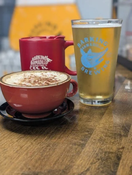 A red coffee cup with a foam-topped cappuccino, a tall glass of beer with a Barking Armadillo Brewing logo, and a red mug with the same logo, all on a wooden table.