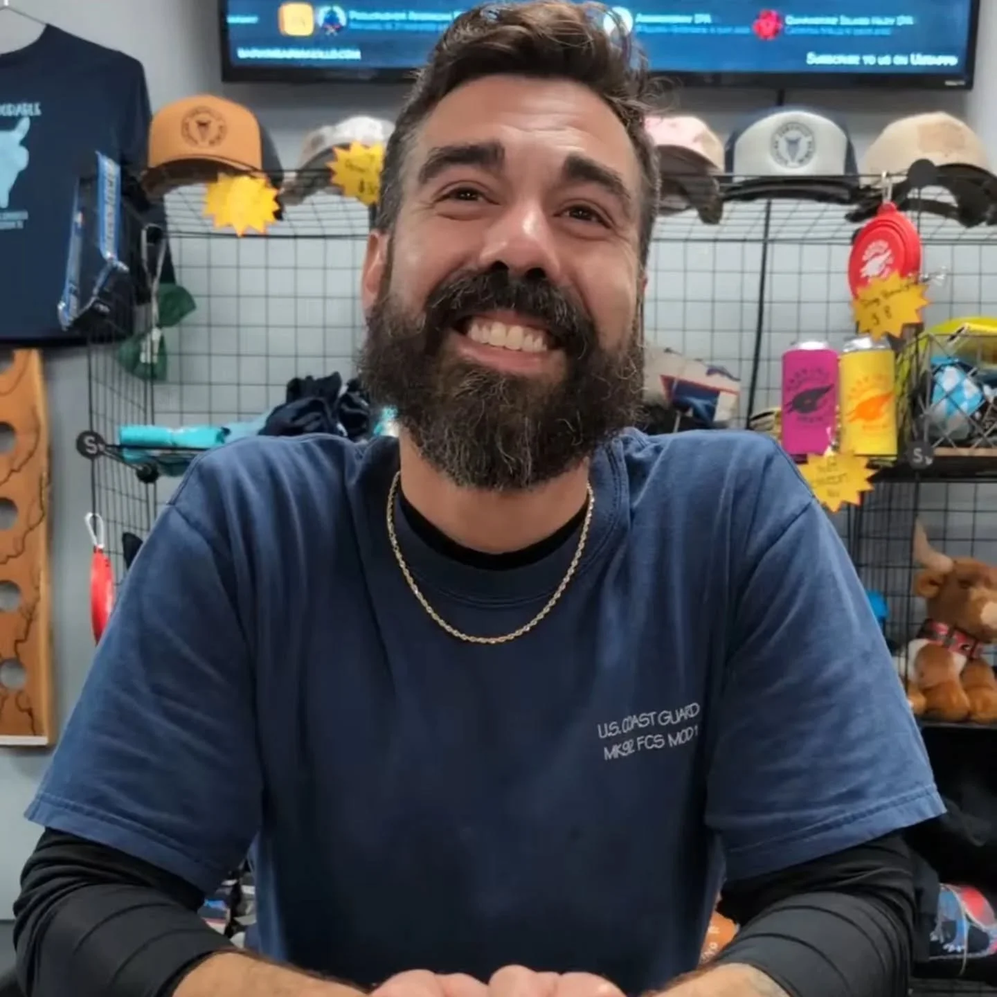 A smiling man with a beard and mustache, wearing a navy blue shirt and gold chains, sitting in front of a display of hats, water bottles, and other merchandise in a store.