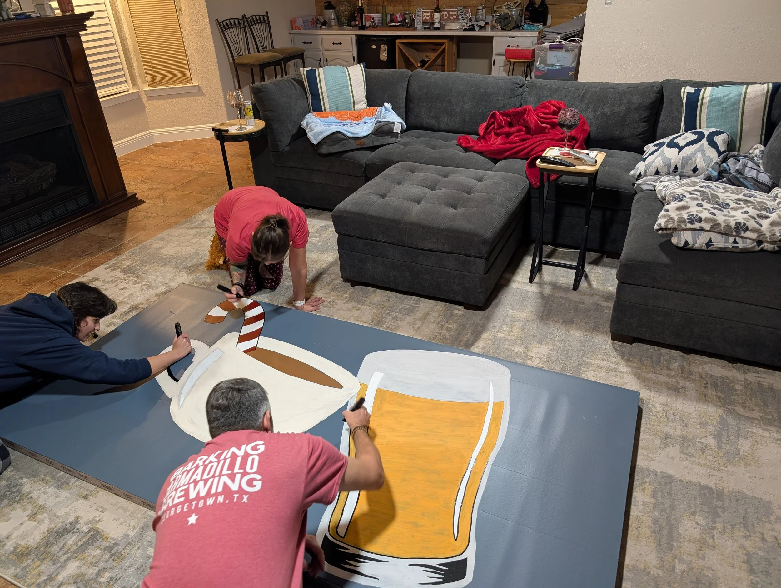 Several people are painting or drawing on a large, flat surface that looks like a wooden cutout of a whale with festive coloring. They are in a living room area with a gray sectional sofa, pillows, blankets, and a side table with a glass of wine. The background includes shelves with bottles and other miscellaneous items.