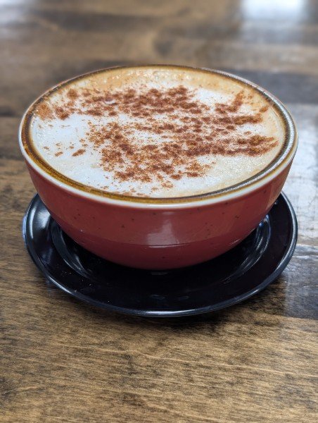 A cup of cappuccino topped with cinnamon or cocoa powder, placed on a black saucer on a wooden table.