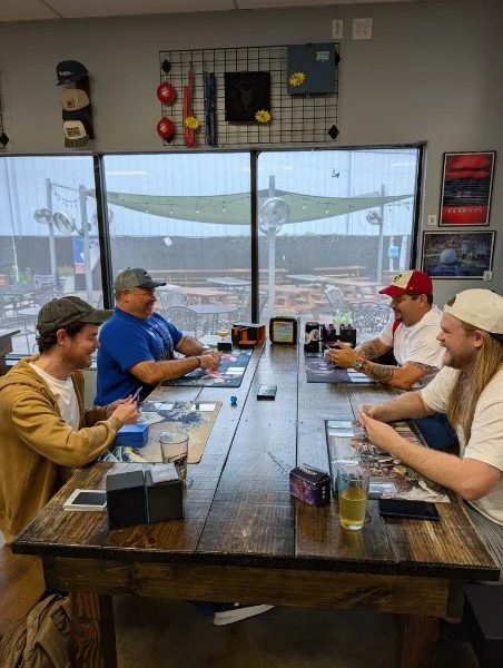 Four men sitting at a wooden table in a restaurant, playing a card game, with drinks in front of them. Large glass windows show an airplane and airport outside.