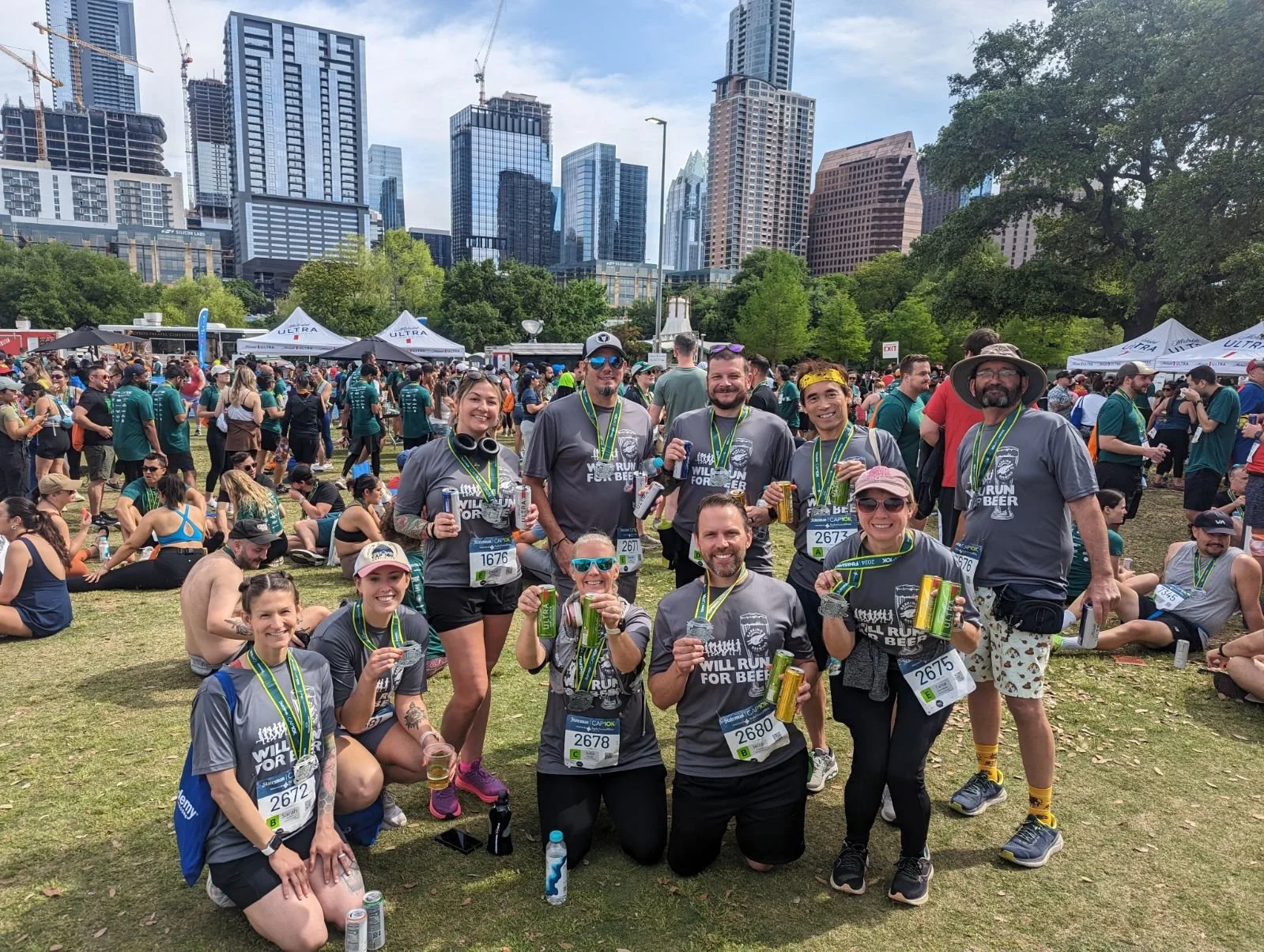 Group of marathon runners smiling, holding drinks, and celebrating at the finish line in an urban park setting with tall city buildings in the background.