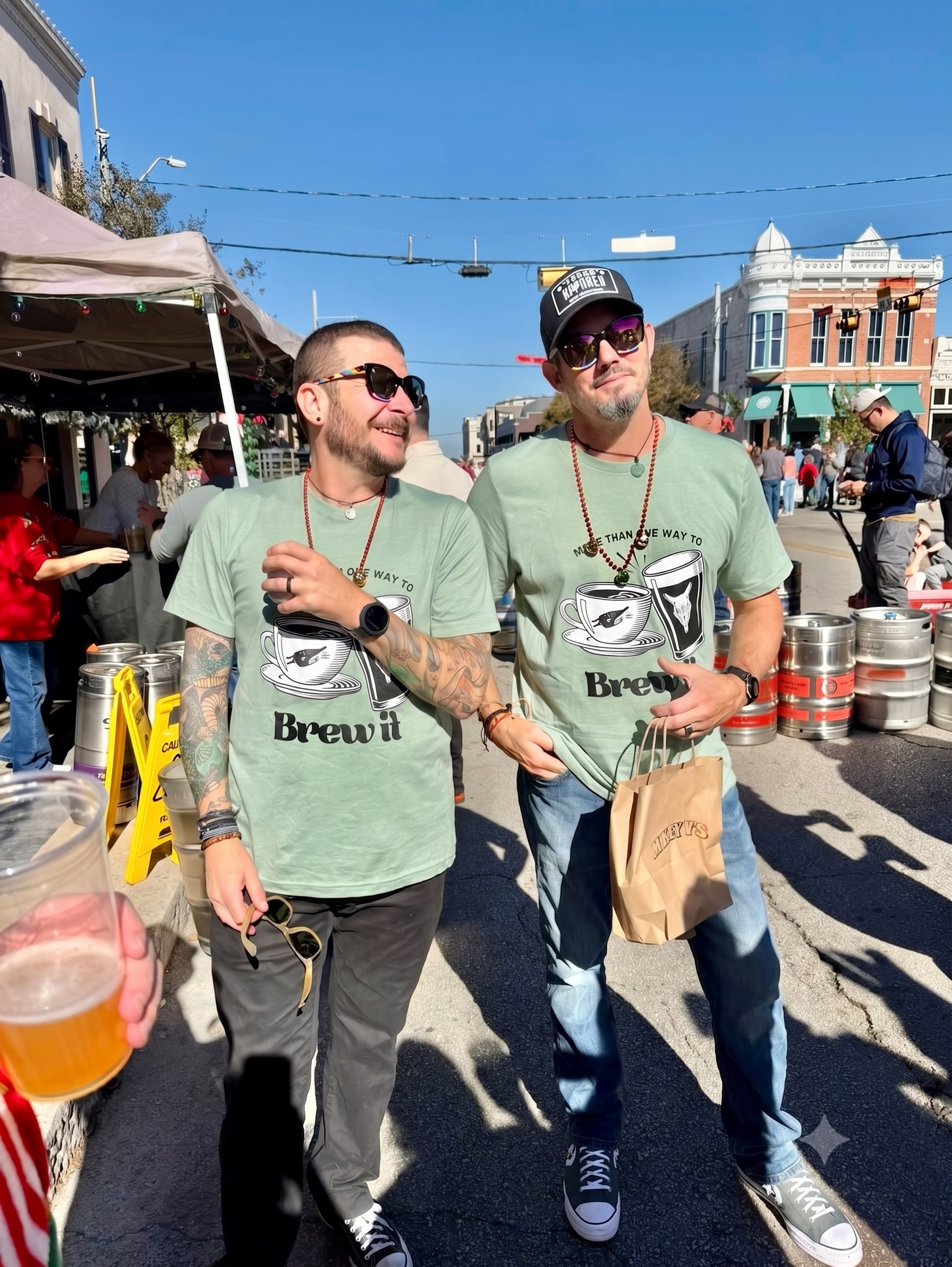 Two men in matching green t-shirts with a coffee design, standing outdoors at an event or festival, with other people, tents, and shops in the background on a sunny day.