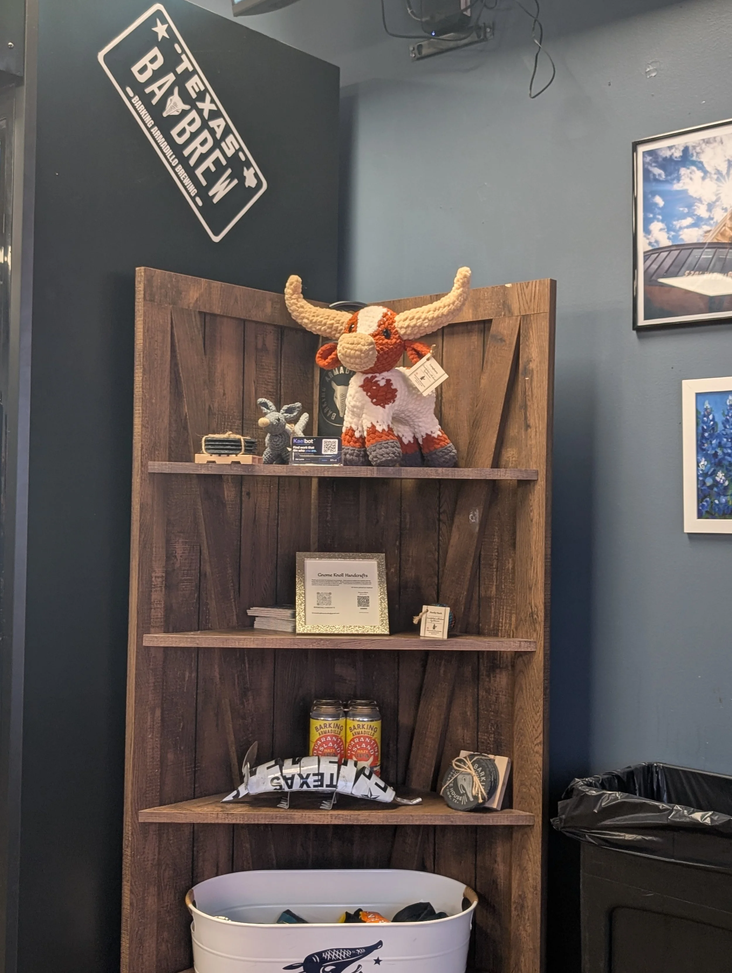 A wooden shelf displaying various items, including a knitted bull toy with large horns, a smaller gray knitted animal, and a framed certificate or flyer. There are also cans labeled 'Barking Armadillo Brewing' on the shelf, and a white container with miscellaneous items inside at the bottom.