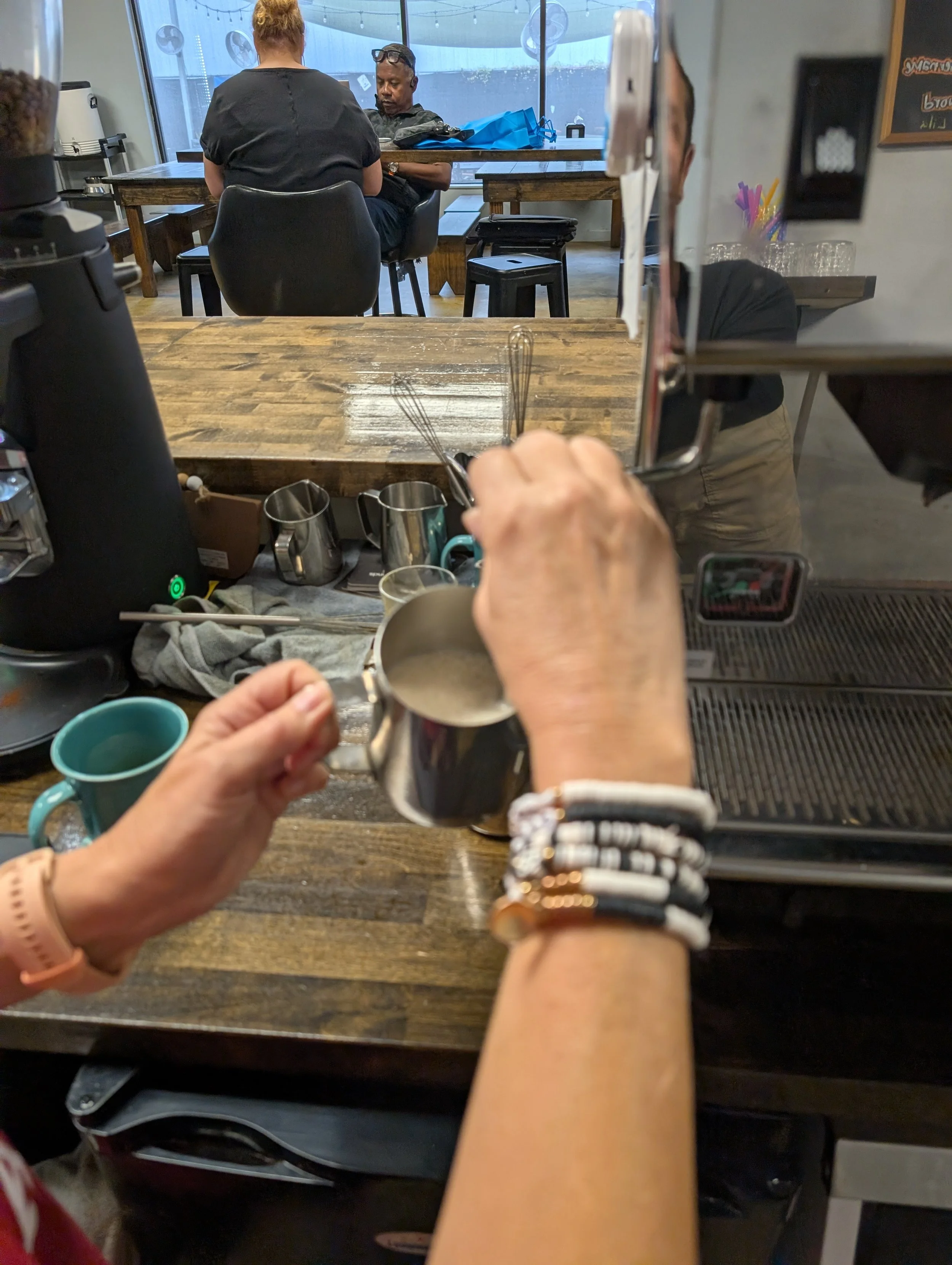 View from behind of a coffee shop counter showing hands holding a stainless steel milk frothing pitcher. On the counter are various coffee-making tools, cups, and a black coffee machine. In the background, two people sit at a table near large windows, one woman and one man, with casual attire.