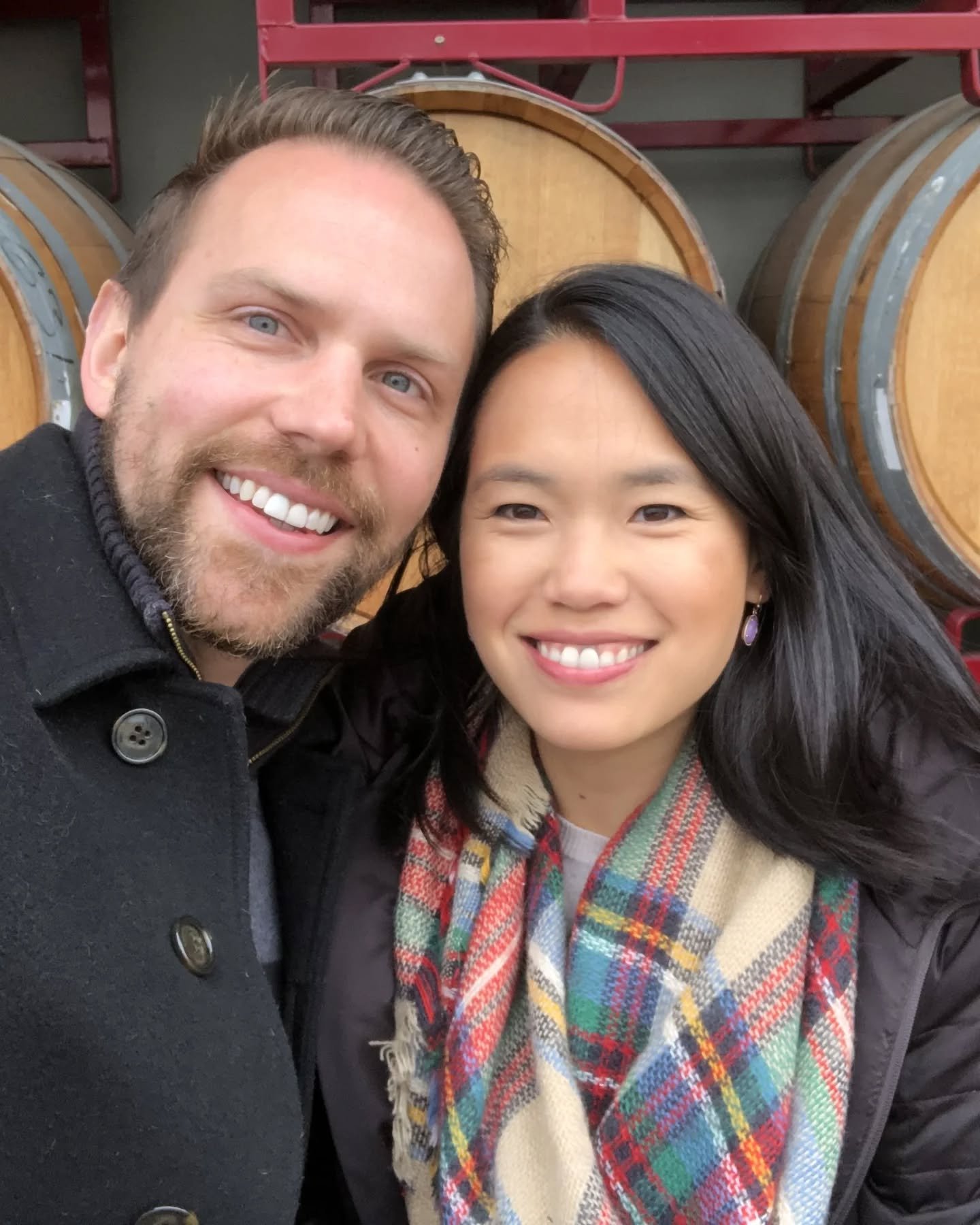 A man and a woman smiling for a selfie in front of stacked wine barrels.