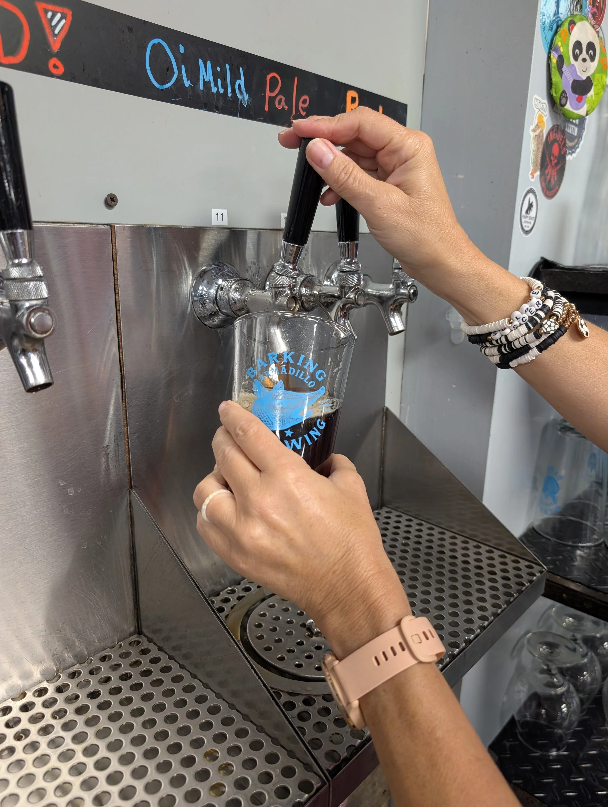 A person is filling a glass with a dark beverage from a tap at a drink station, with a sign indicating the beverage options labeled "O (Mild)" and "Pale." The person has a beige wristwatch and several black and white beaded bracelets.