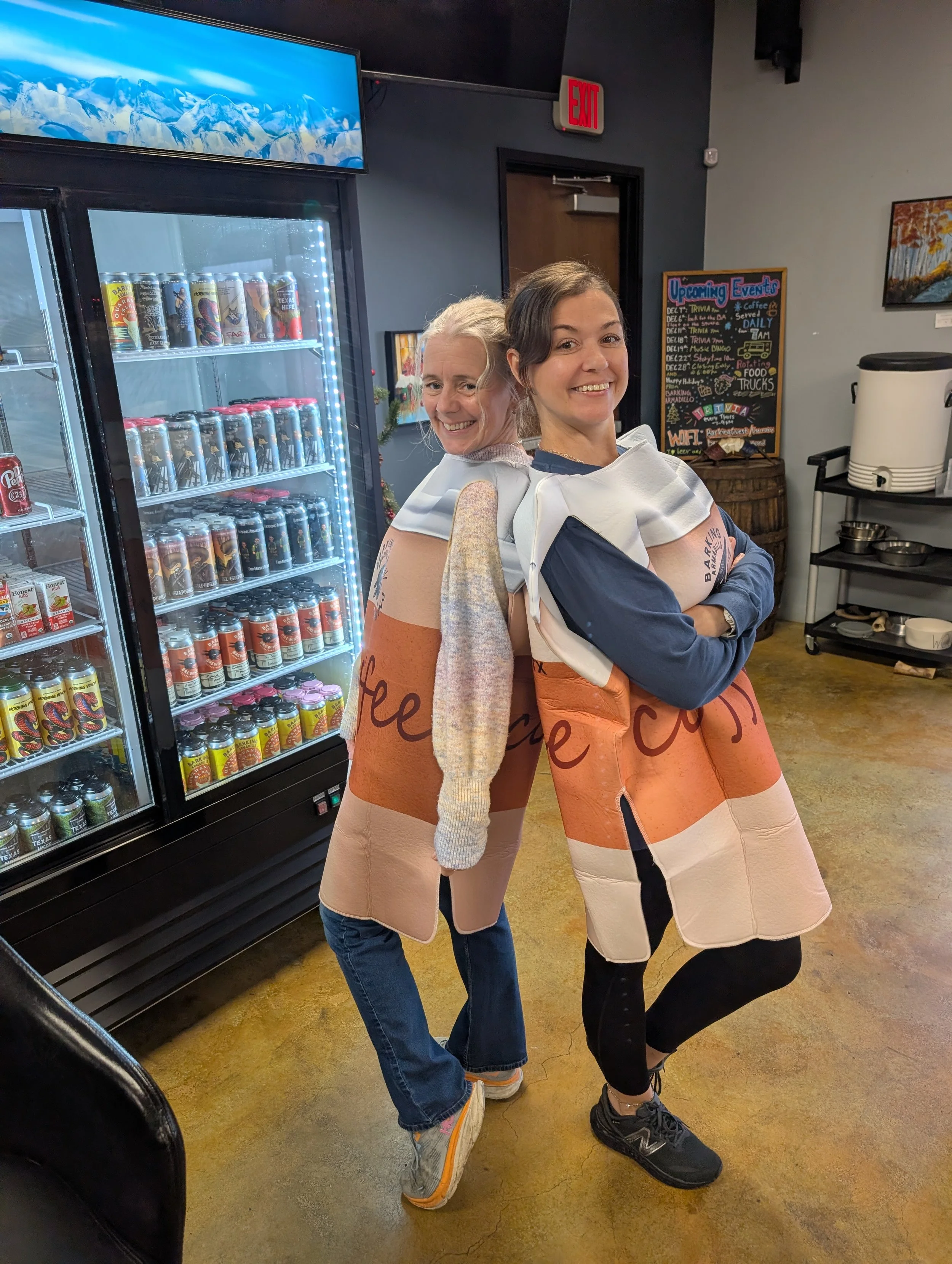 Two women standing back to back wearing large costumes made from cardboard boxes designed to look like beer cans, inside a store near a refrigerator filled with canned drinks and a chalkboard event sign.