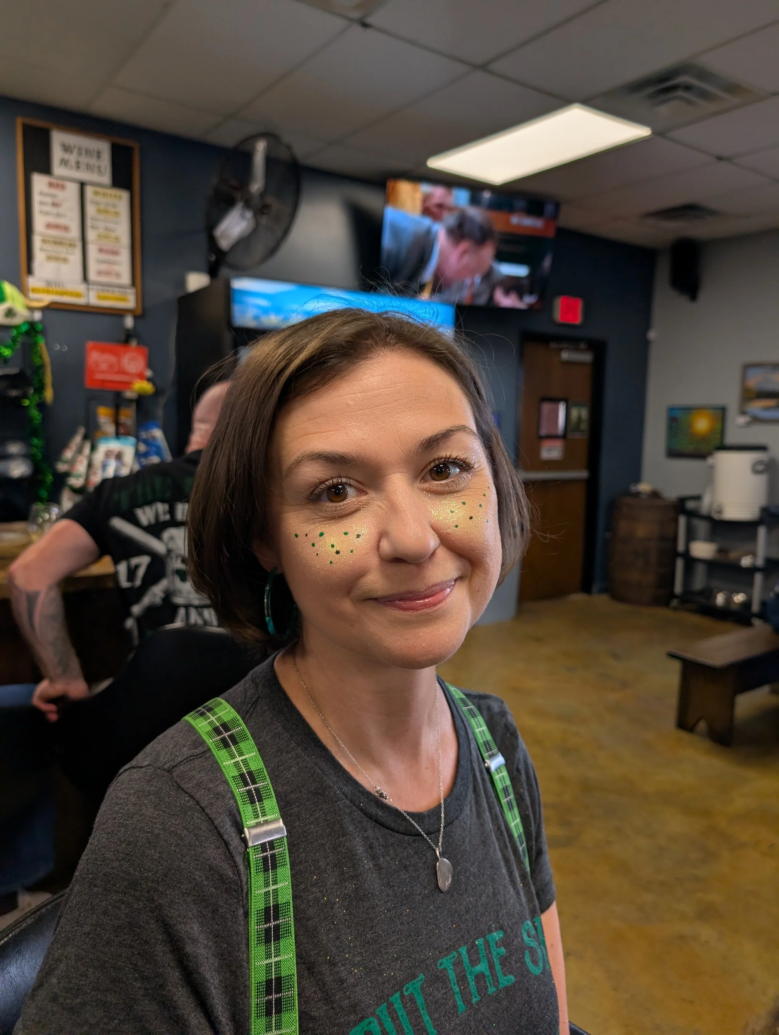 A woman with short brown hair, green polka dot face decorations, and a silver necklace, smiling, wearing a black shirt with green plaid straps, in a room with a television showing two men in a conversation.