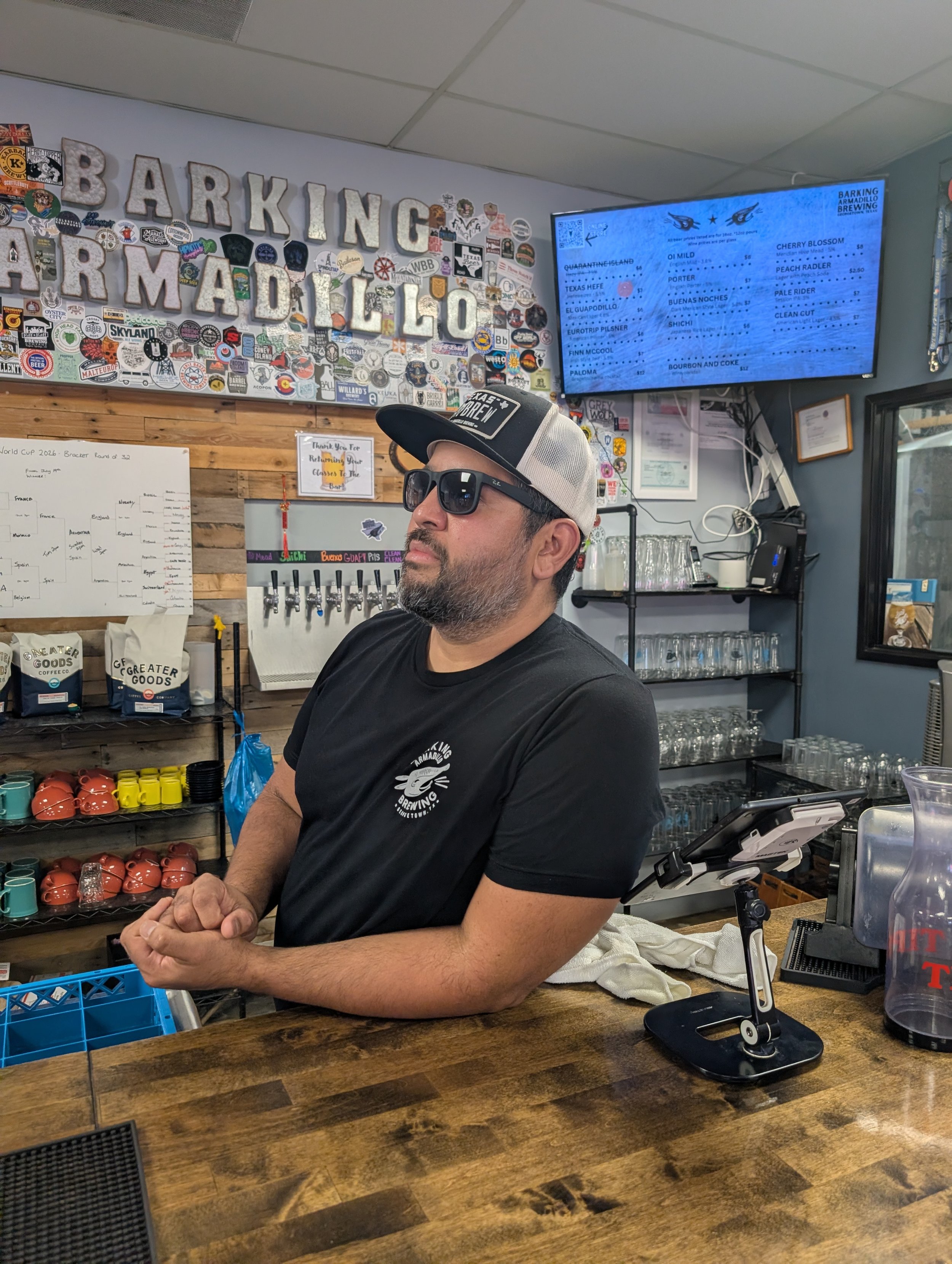A man wearing a black T-shirt, sunglasses, and a trucker hat, sitting at a wooden counter in a bar or brewery. Behind him, there are shelves with coffee mugs, glasses, and bags of coffee beans, and a digital menu screen displaying drink options. The background wall is decorated with numerous brewery and beer stickers and signs.