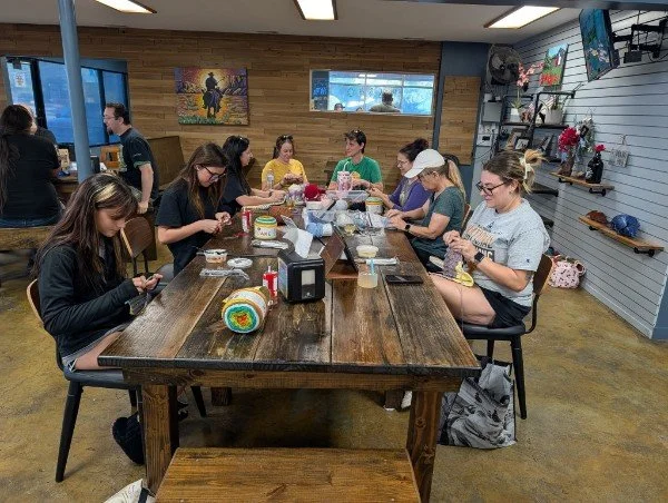A group of women sitting at a long wooden table in a craft store, working on arts and crafts projects.