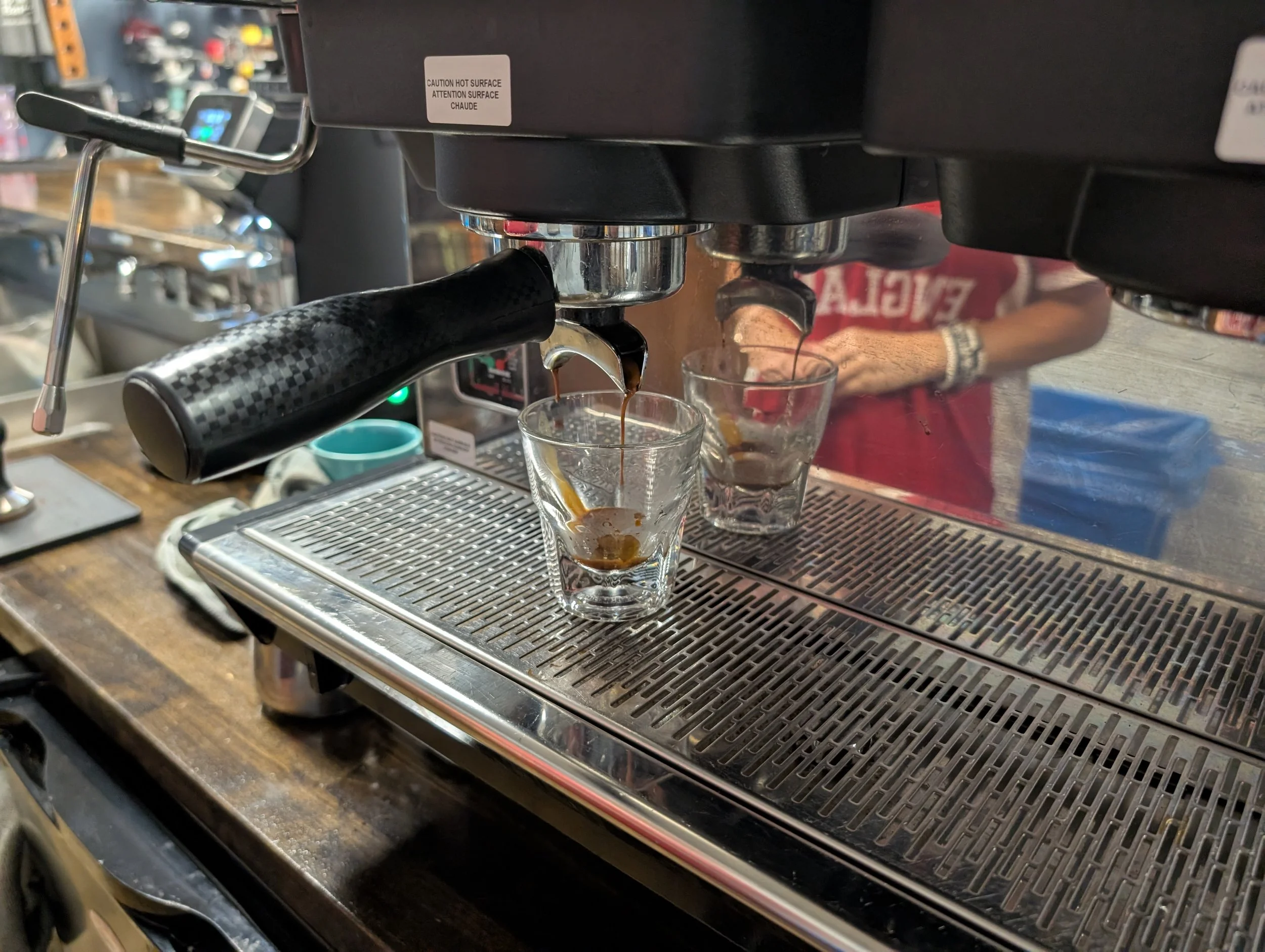 Close-up of a coffee machine dispensing coffee into glasses, with a person in the background wearing a red shirt.