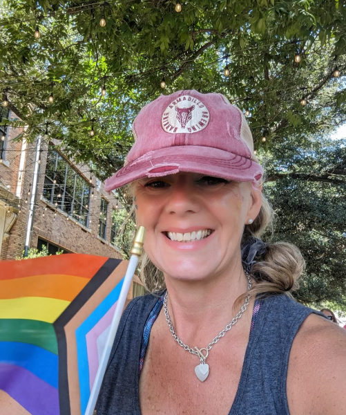 A woman smiling outdoors, wearing a pink cap, holding a rainbow pride flag, with trees and string lights in the background.
